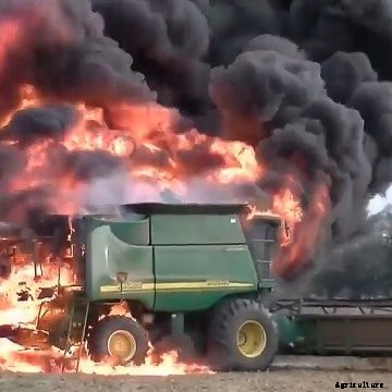 A John Deere combine engulfed in flames sits in a soybean field with black smoke billowing