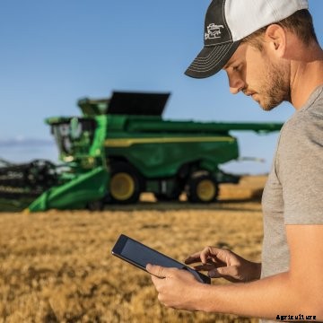 A man stands in a field with an iPad with a green John Deere combine behind him